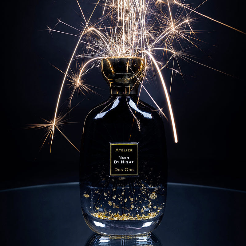 Noir By Night Atelier Des Ors perfume bottle with gold flakes and sparkling fireworks background