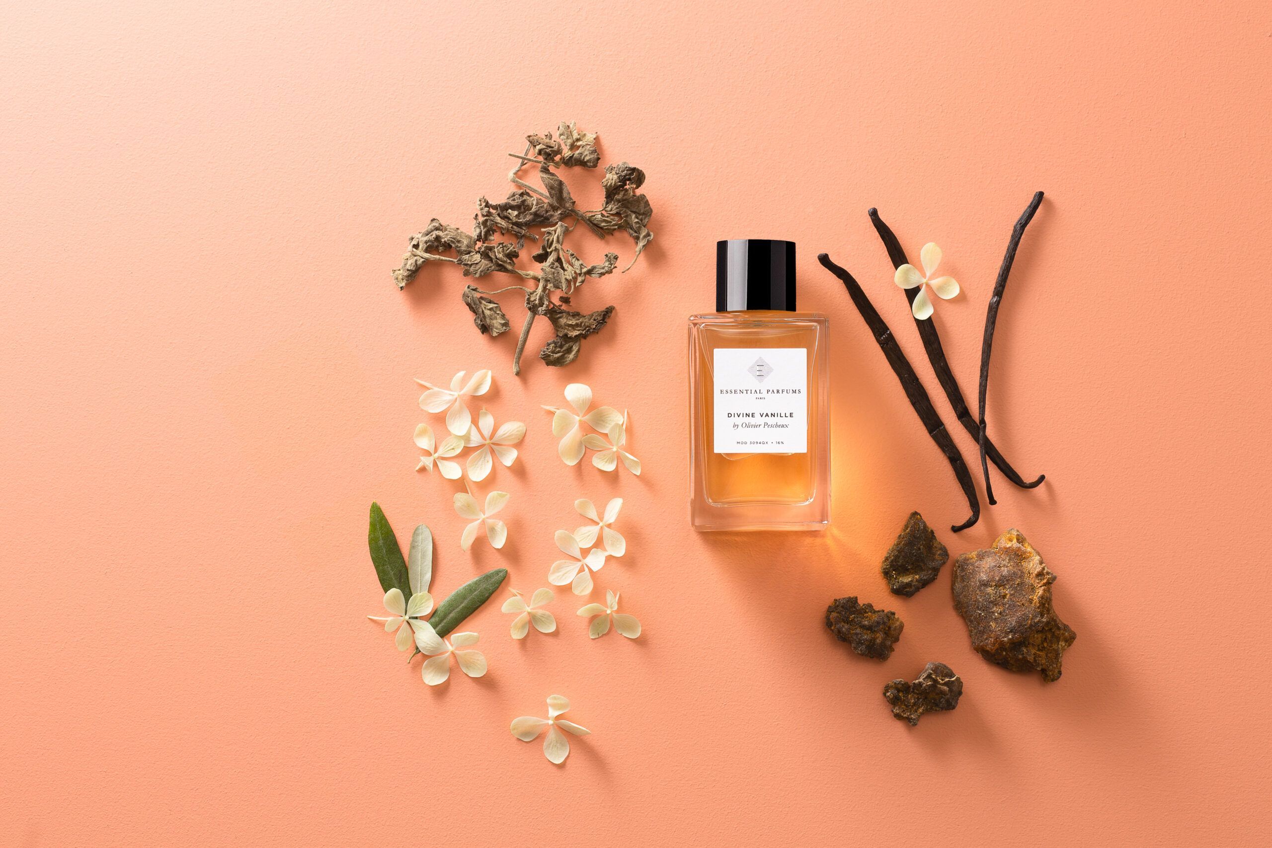 Essential Parfums Divine Vanille eau de parfum bottle surrounded by vanilla pods and floral ingredients on peach background