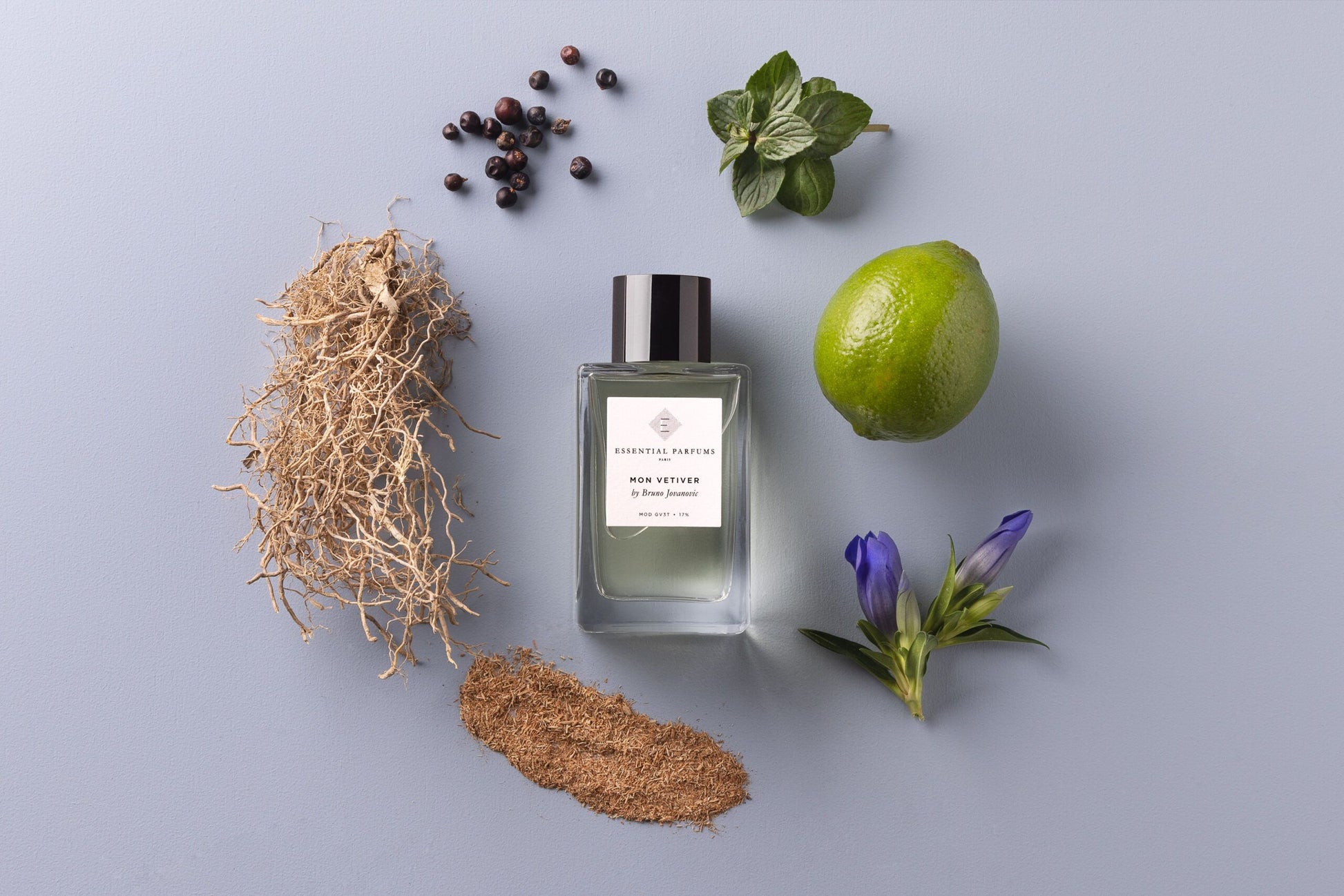 Essential Parfums Mon Vetiver 100ml perfume bottle surrounded by lime, roots, green leaves, blue flowers, and black peppercorns