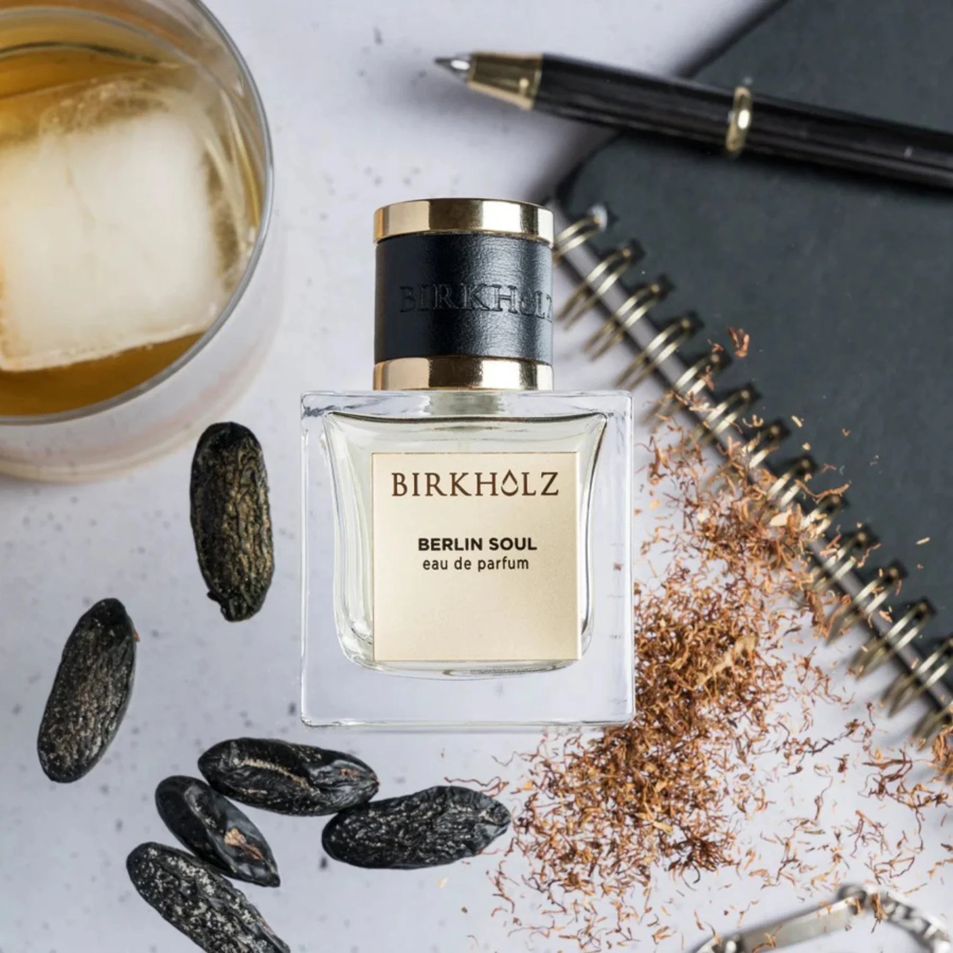 Birkholz Berlin Soul niche perfume bottle with black cap beside spices, notebook, pen, and glass of iced drink