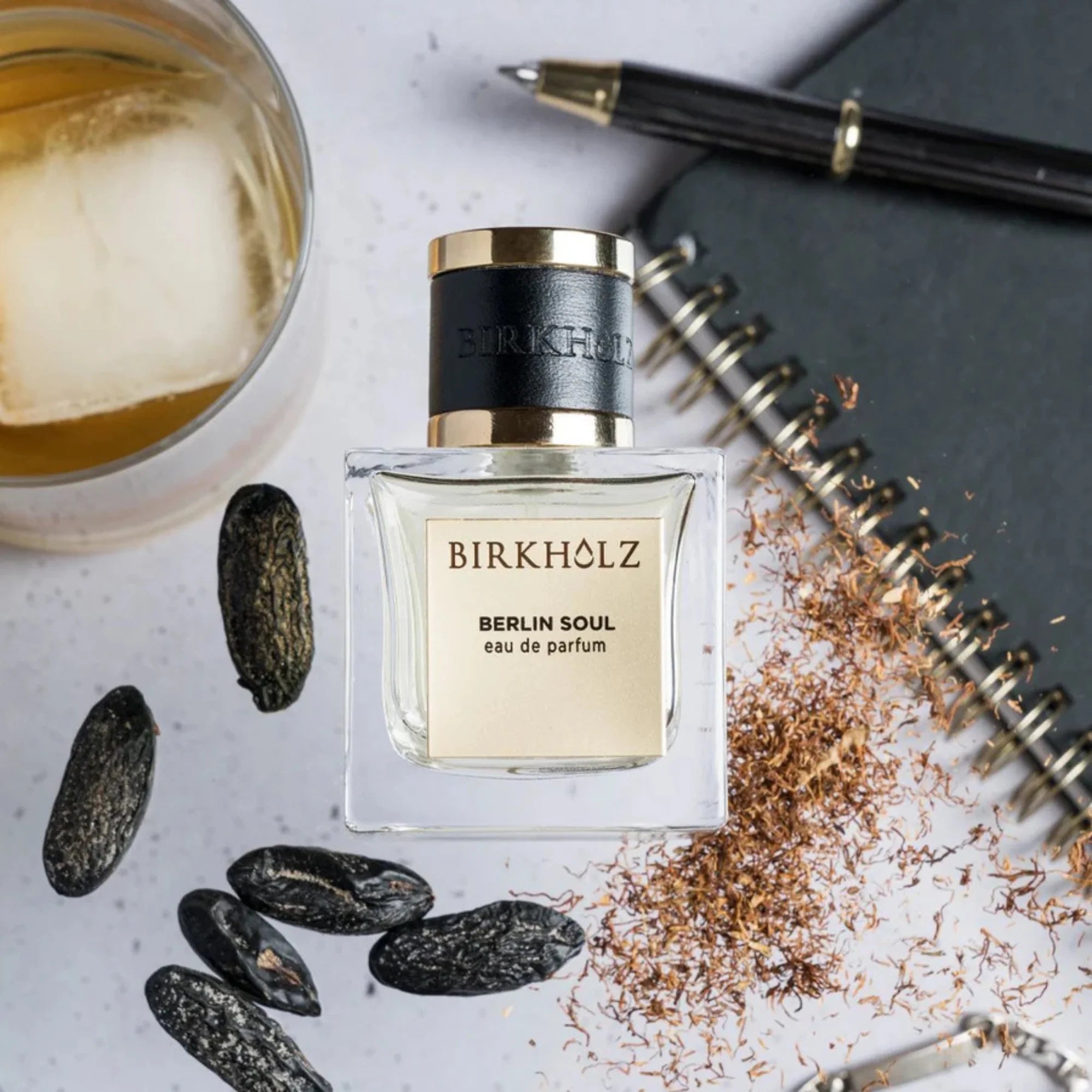 Birkholz Berlin Soul niche perfume bottle with black cap beside spices, notebook, pen, and glass of iced drink