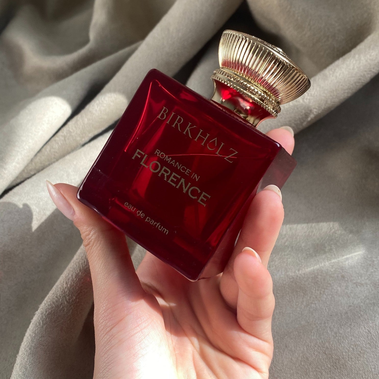 Birkholz Romance in Florence perfume bottle in deep red with gold cap held in hand on a soft fabric background