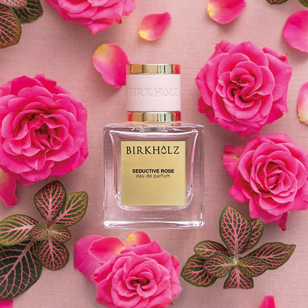 Birkholz seductive rose perfume bottle surrounded by vibrant pink roses and green leaves on soft pink background