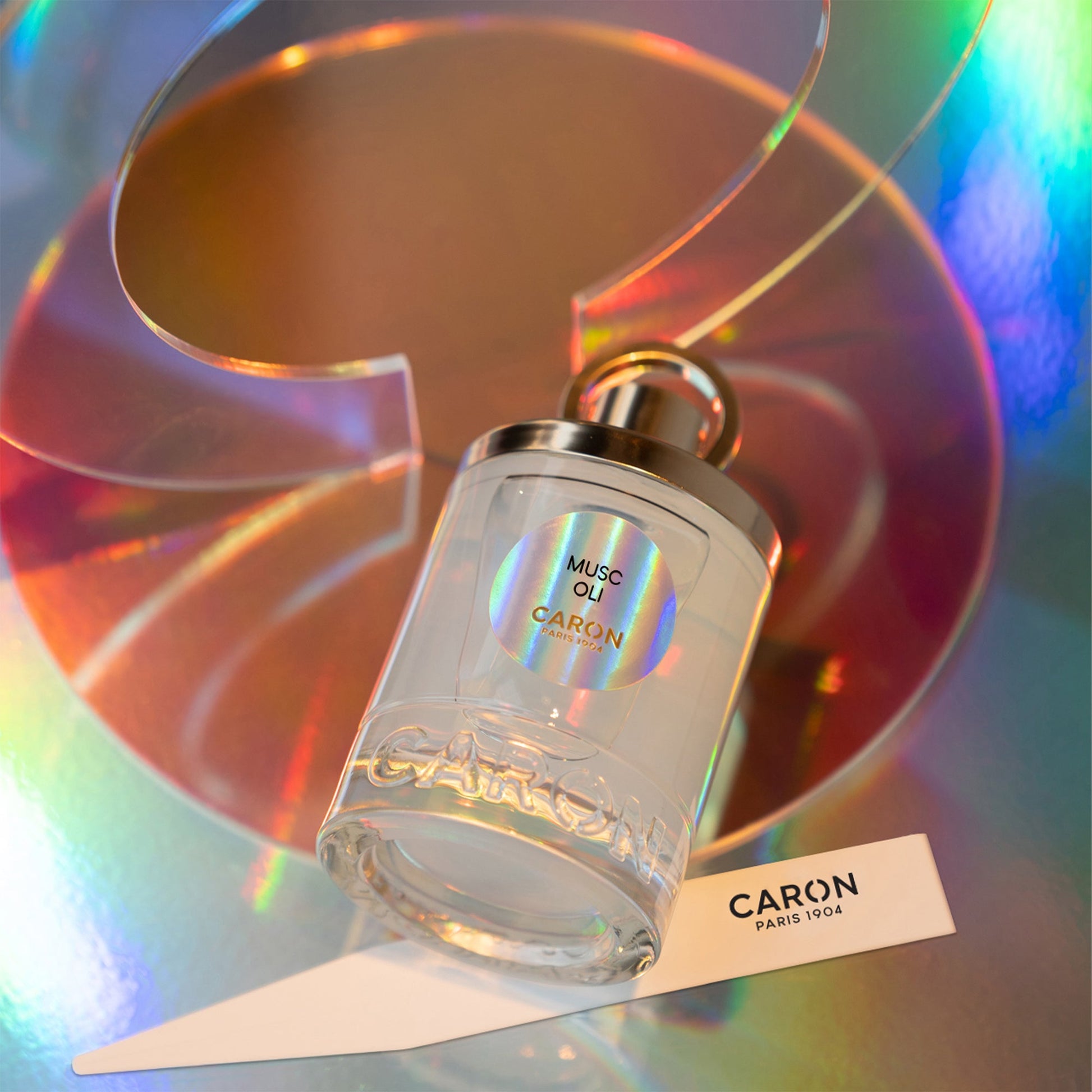 Caron Musc Oli Eau De Toilette clear glass perfume bottle with silver cap on reflective colorful background