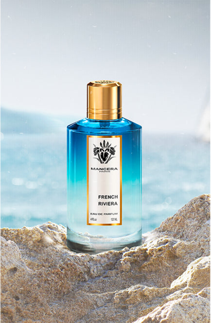 Mancera French Riviera Eau de Parfum bottle with blue gradient glass and gold cap on rocky beach background