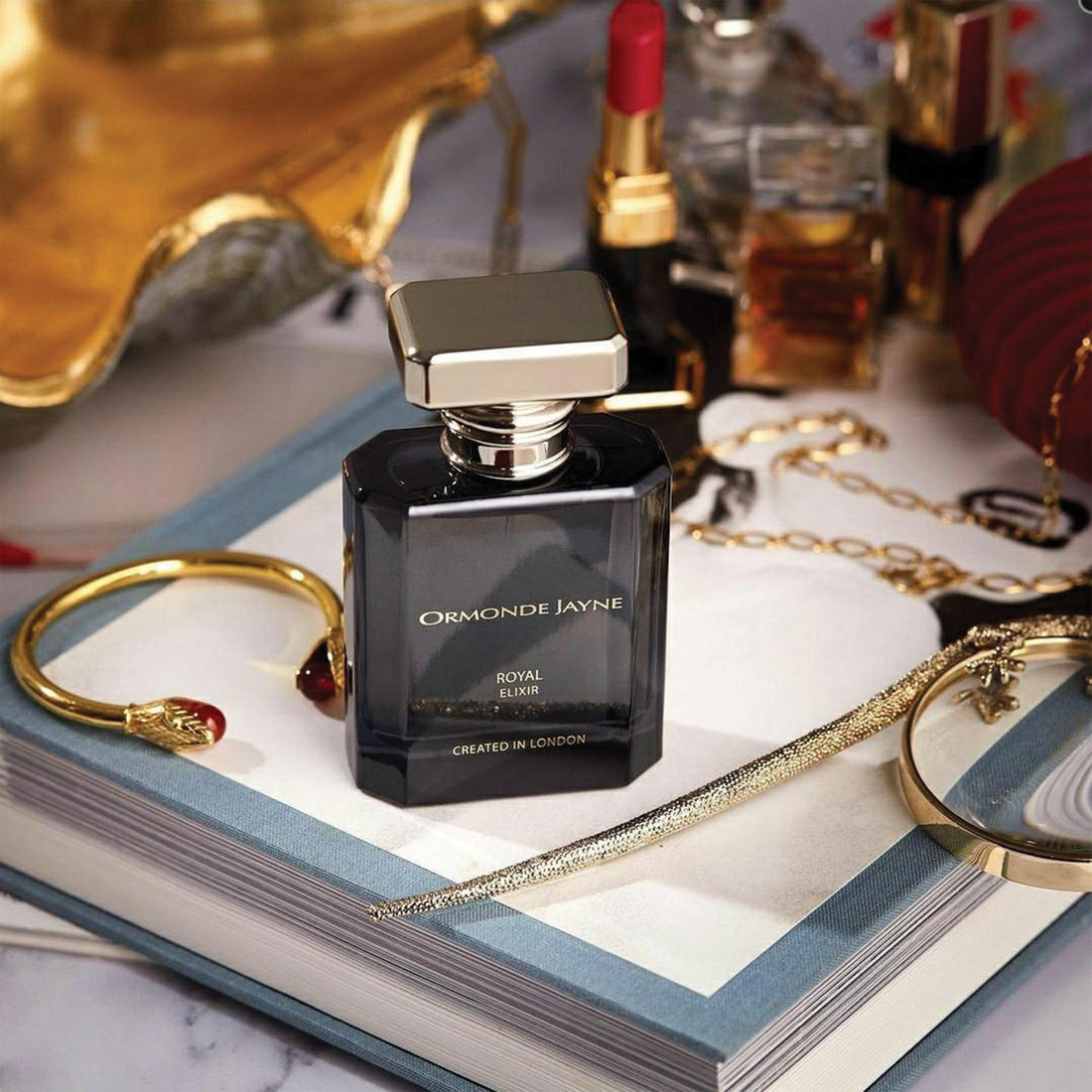 Ormonde Jayne Royal Elixir fragrance 50ml bottle placed on an open book with gold jewelry and lipstick nearby