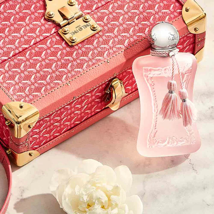 Parfums De Marly Delina La Rosee perfume bottle with pink tassels next to a pink textured suitcase and white flower