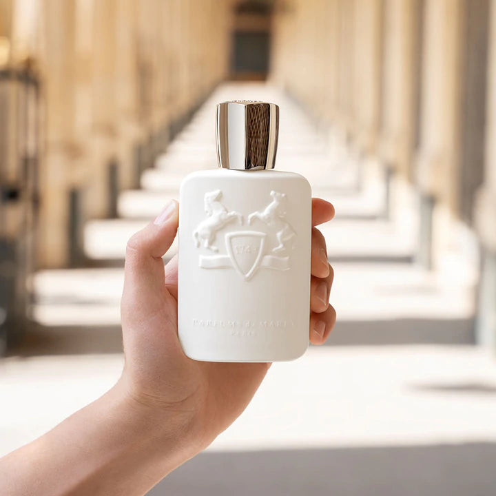 Hand holding Parfums De Marly Galloway Eau de Parfum white bottle with silver cap in blurred outdoor setting